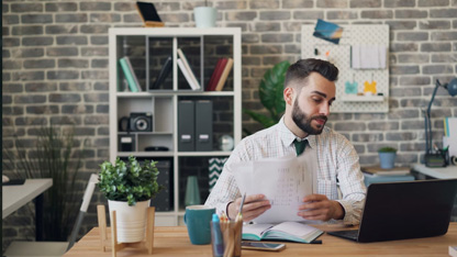 Un bărbat care lucrează la un birou cu un laptop, ținând documente, într-un birou modern cu pereți din cărămidă.